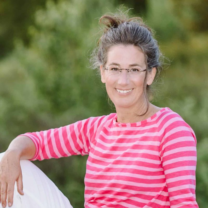Claudia Drescher sitzt im Wald und hält einen Arm über ihr Knie und lacht.